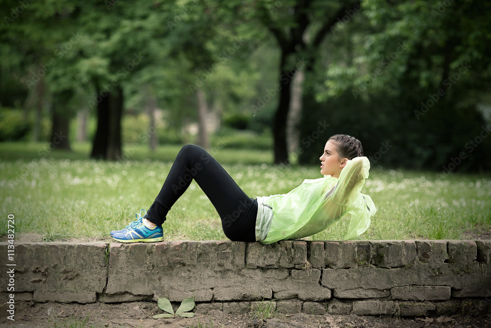Side view of a young woman exercising outdoors. She is sitting on the wall and doing sit-ups.