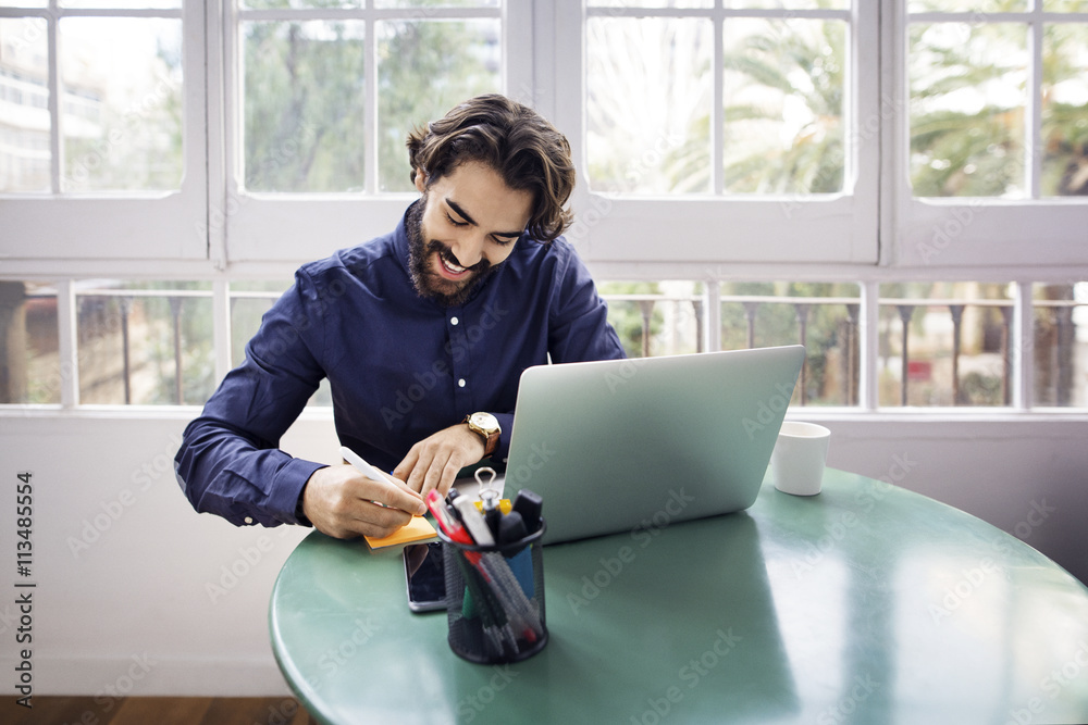 © Cavan Images - Happy businessman writing on adhesive note while sitting at table with laptop in office