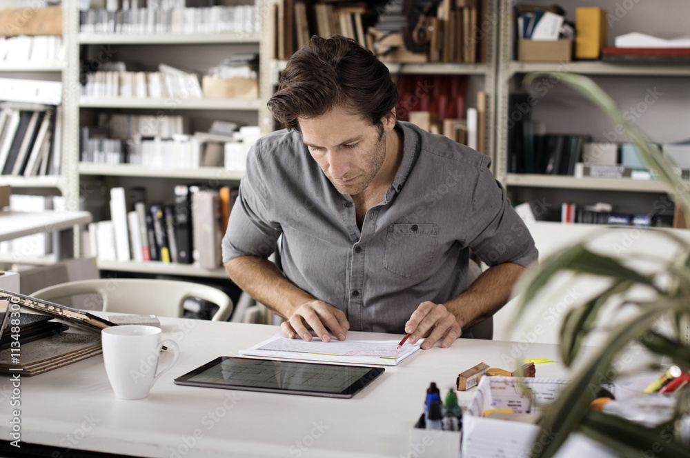 Male interior designer using tablet computer while analyzing documents at desk in office Male interior designer using tablet computer while analyzing documents at desk in office