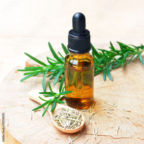a bottle of rosemary aromatherapy oil extract with fresh and dried rosemary leaf on wooden spoon. 