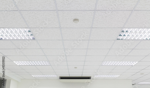 Office ceiling in perspective with white texture of acoustic gypsum plasterboard, lighting fixtures or fluorescent panel light suspended on square grid structure. Interior modern building background.