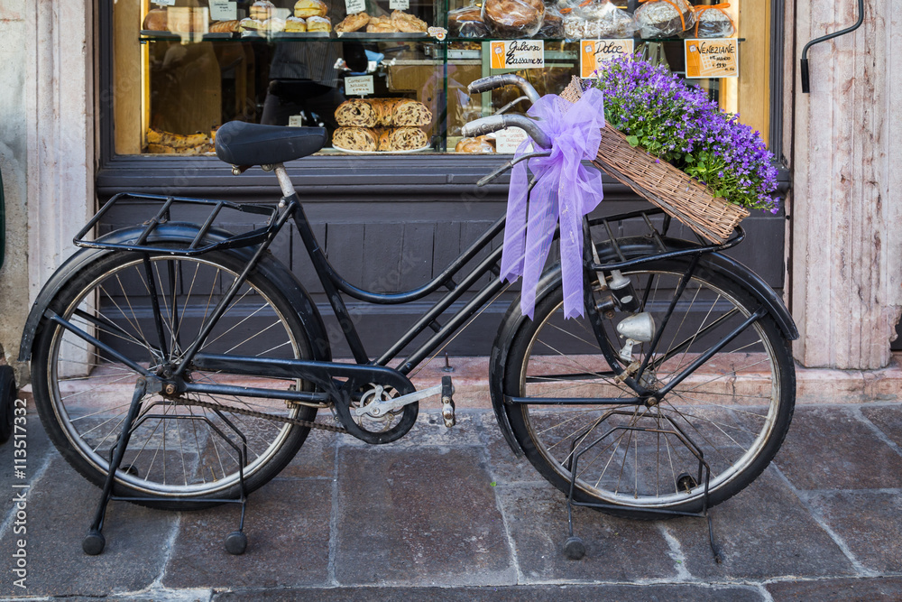 Black old bicycle decorated with purple flower basket and a gift wrap, infront of a backery in Bassano del Grappa, Italy