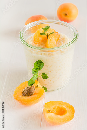 Ein Glas frisch gekochter Milchreis mit Aprikosen steht auf einem weißen Tisch.