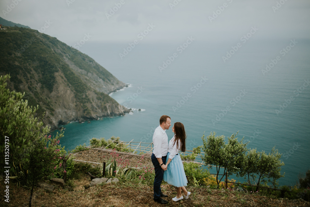 Young cute couple honeymoon posing and holding their hands on dating in a beautiful place italy near ocean and mountains, hug, smile and talk to each other

