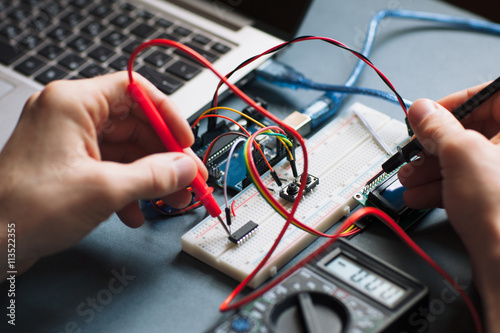 Single board microcontroller testing close-up. Electrical tester units in engineer hands, working with microcontroller, connected to laptop on black background
