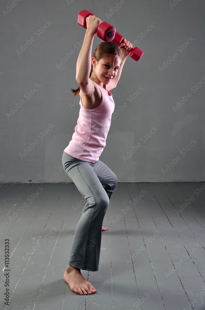Teenager girl doing exercises with dumbbells to develop with dumbbells muscles on grey background. Full length portrait of teen child exercising with weights.