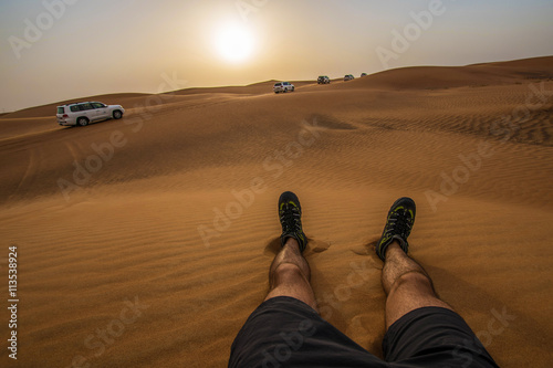 Watching sunset in Dubai sitting on dunes - point of view legs and safari jeeps