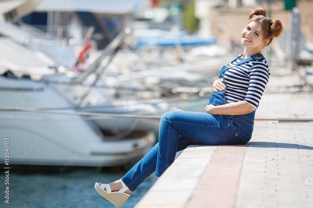 Stylish pregnant brunette woman with trendy hairstyle,dressed in blue denim overalls and striped t-shirt,wearing jewelry posing on the promenade,sitting on the dock in front of the yachts and boats