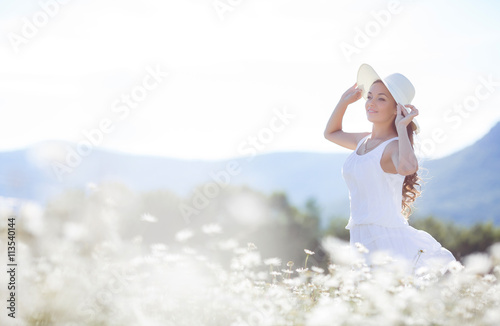 Portrait of beautiful young woman in a white hat with a wide brim,brunette with long curly hair in white summer dress posing in a field of blooming white daisies in a mountain area