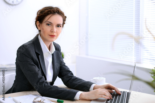 Business woman with a cup of coffee is sitting at the table and typing on a laptop computer in the white colored office . Ukrainian hair styling. Independence and success concept.