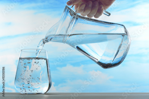 Fresh and clean drinking water being poured from jug into glass on sky background
