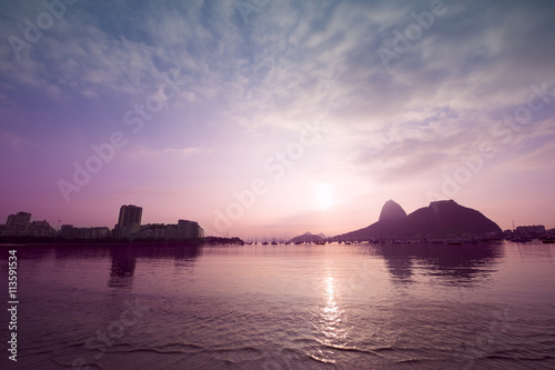 Colorful sunrise reflects on the calm surface of Botafogo Bay with a silhouette of Sugarloaf Mountain in Rio de Janeiro, Brazil © lazyllama