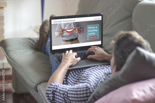Young man lying comfortably on the sofa while using the laptop with dental insurance plan on screen. View from behind. All screen graphics are made up.