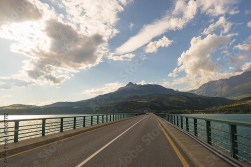 Paved two lane road on bridge crossing lake in scenic landscape and moody sky. Panoramic view from car mounted camera. Summer adventure and roadtrip in vacations.