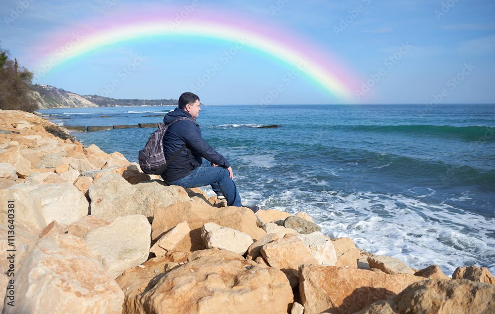 Young male traveler sitting on the rocks and enjoy the views of the sea and nature rainbow