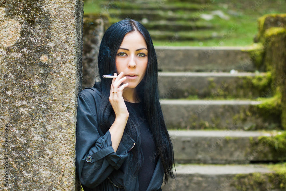 Beautiful girl in black coat and dress standing next to wall on old stairs in the city, grass and trees in background, smoking a cigarette Beautiful girl in black coat and dress standing next to wall on old stairs in the city, grass and trees in background, smoking a cigarette