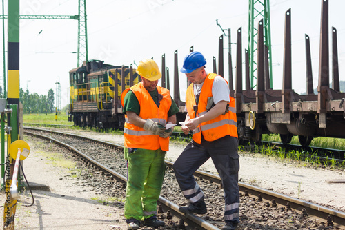 Rail workers coordinating cargo loading to transport train on the tracks. Railway employees in safety west and helmet talking about instructions.
