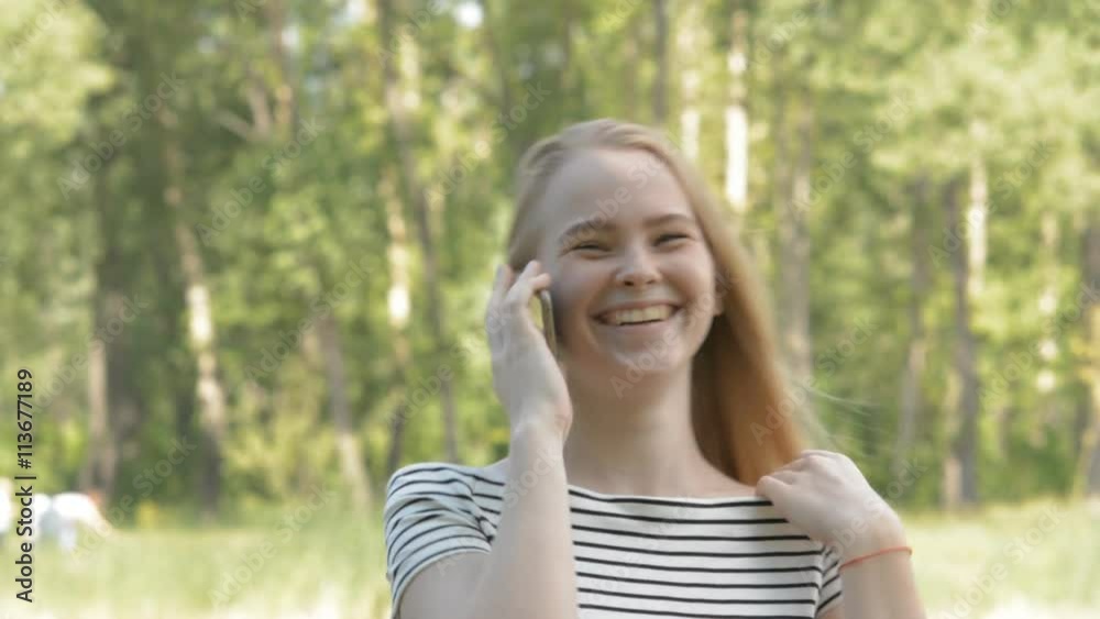 Closeup of a cheerful young woman talking on the phone outdoors in a park. Girl with a cell phone on a green background of blurred trees. Closeup of a cheerful young woman talking on the phone outdoors in a park. Girl with a cell phone on a green background of blurred trees.