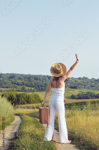 Backview of female holding retro suitcase on the blue sky sunny outdoors country road background.