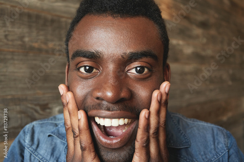 Happy and amazed African American male looking exuberantly at camera with hands on cheeks and mouth opened. Attractive man in denim short on brown background agreeably surprised by good news. © wayhome.studio 