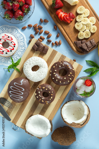Delicious donuts. Served on the wooden board and white napkin. Decorated with strawberry, banana, coconut, mint and almond. Blue background. Vertical image. With lemonade. Top view