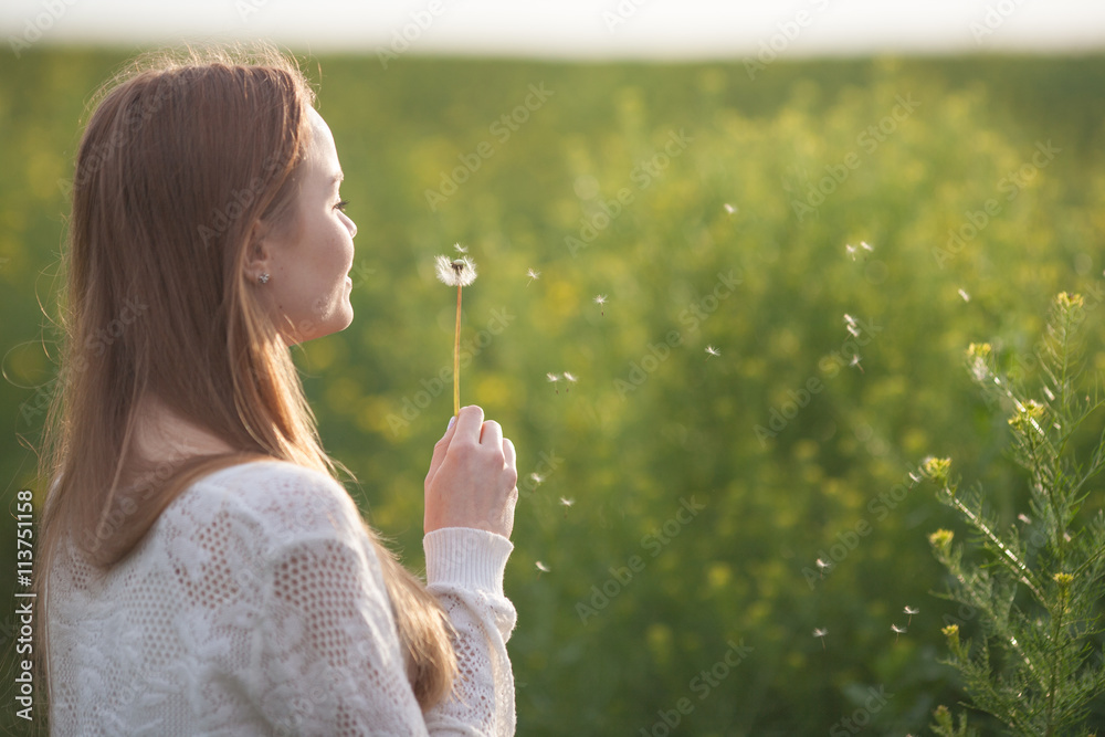 Young spring fashion woman blowing dandelion in spring garden. Springtime. Trendy girl at sunset in spring landscape background. Allergic to pollen of flowers. Spring allergy.