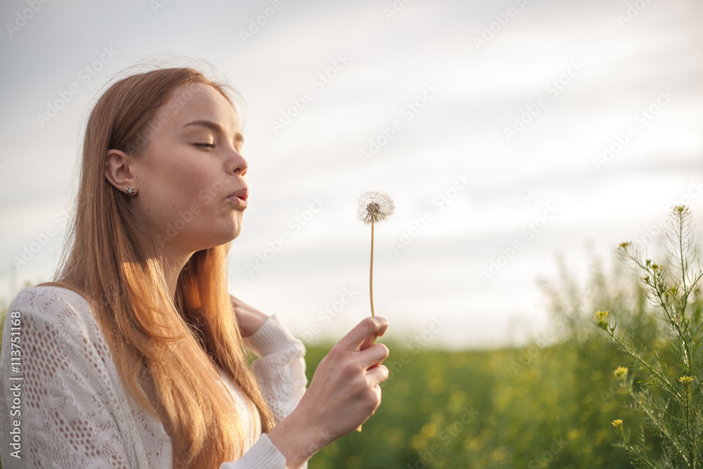 Young spring fashion woman blowing dandelion in spring garden. Springtime. Trendy girl at sunset in spring landscape background. Allergic to pollen of flowers. Spring allergy.
