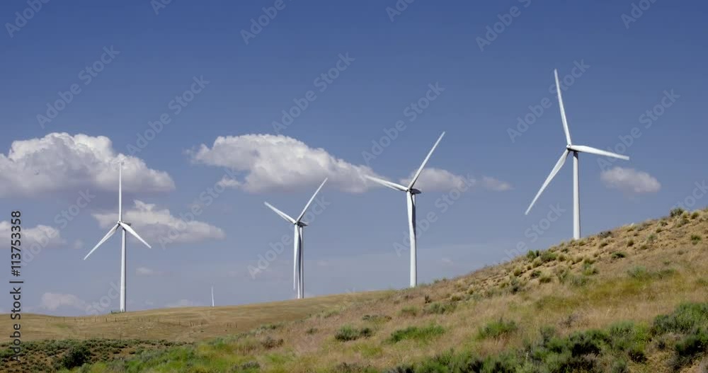 Tilt down from blue sky to large wind turbines operating on a rural hillside in the Tehachapi Mountains in California. Blue sky and puffy cumulus clouds behind them. Tilt down from blue sky to large wind turbines operating on a rural hillside in the Tehachapi Mountains in California. Blue sky and puffy cumulus clouds behind them.
