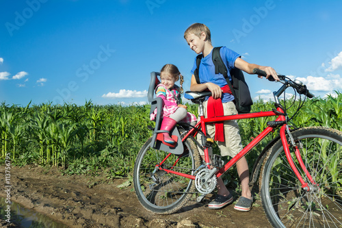 Sibling teenage boy cycling with his little sibling sister on baby bike seat on farm corn field dirt road
