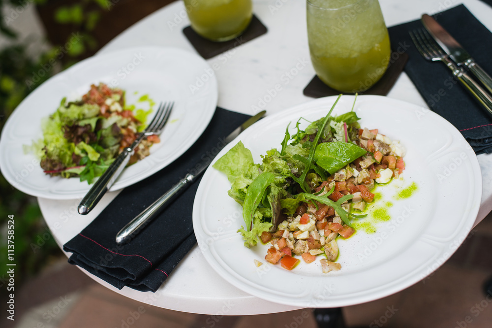 Two plates of fresh salad with arugula, mozzarella, vegetables, tomatoes, chicken served with mint lemonade in a small outdoor restaurant at the lunch