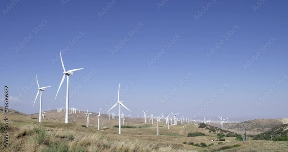 Tilt down from blue sky to wide view of wind turbines operating on rural hillsides in the Tehachapi Mountains in California.  Blue sky and a house can be seen behind them.