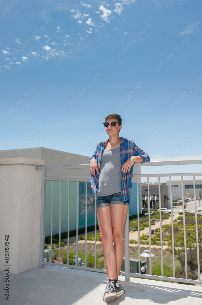 Teenage girl with crew cut leaning back on handrail under blue sky.