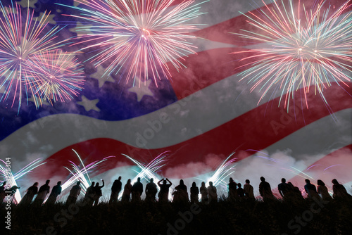 Group of people enjoying fireworks show in a carnival or holiday. People in silhouette and blurred American flag in background.