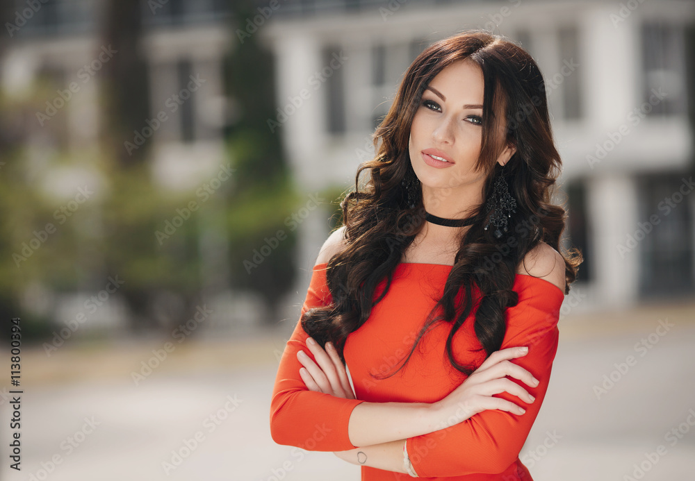 Summer portrait of a beautiful,brunette with long curly hair and grey eyes, dressed in a bright orange dress,wearing large black earrings,a cute smile posing outdoors in the city in the summer
