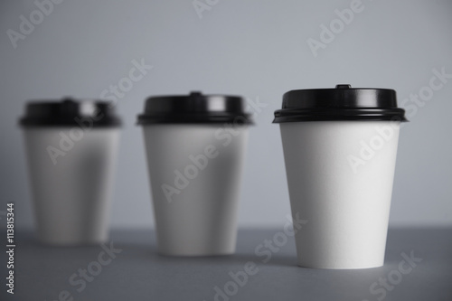 Three take away white paper cups with closed black caps, top view, isolated on simple gray background, first cup in close focus, cups behind are unfocused in bokeh