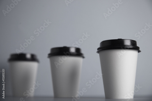 Three take away white paper cups with closed black caps, top view, isolated on simple gray background, first cup in close focus, cups behind are unfocused , bottom view