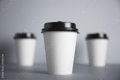 Three take away white paper cups with closed black caps, top view, isolated on center of simple gray background, first cup in close focus, cups behind are unfocused in bokeh