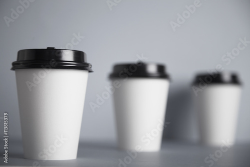 Three take away white paper cups with closed black caps, top view, isolated on simple gray background, left cup in close focus, cups behind are unfocused in bokeh