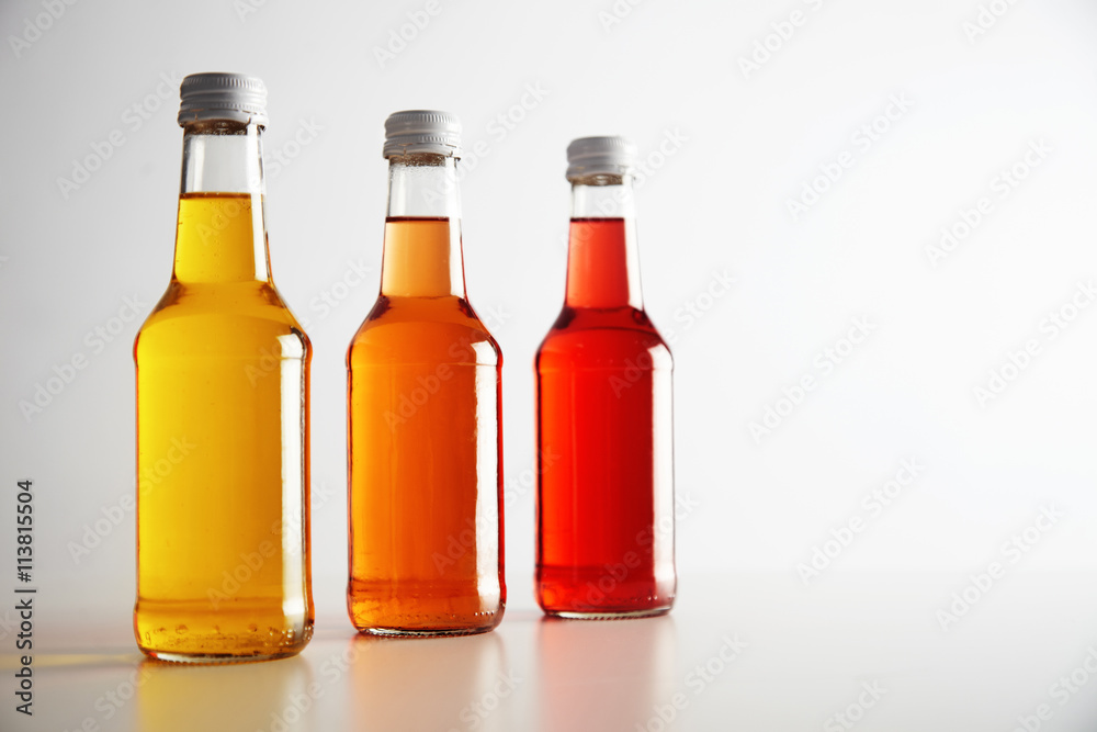 Three glass bottless with colored drinks inside: red, orange, yellow. Presented on white table, isolated on white. Ready for labeling and sale