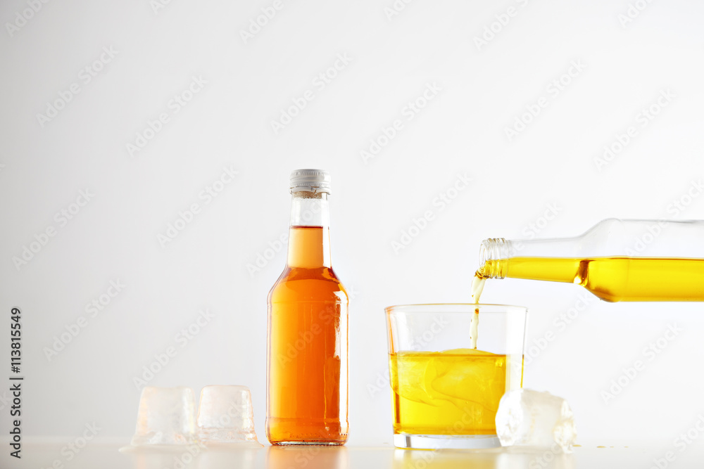 Yellow tasty lemonade drink pours from bottle to glass with ice cubes near closed sealed unlabeled bottle with orange beverage, isolated on white Yellow tasty lemonade drink pours from bottle to glass with ice cubes near closed sealed unlabeled bottle with orange beverage, isolated on white