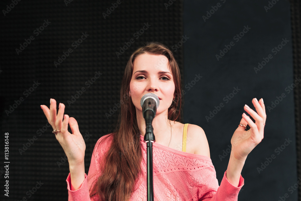 Fototapeta premium Young singing woman inro the microphone close-up. Beautiful woman in pink dress sings into the micriphone, hands are rised up. Songing woman looking at camera