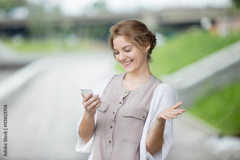 Portrait of smiling beautiful caucasian woman walking on the street and looking at smartphone screen with joyful expression. Casual female laughing at texts or video on phone outdoors in summer Portrait of smiling beautiful caucasian woman walking on the street and looking at smartphone screen with joyful expression. Casual female laughing at texts or video on phone outdoors in summer