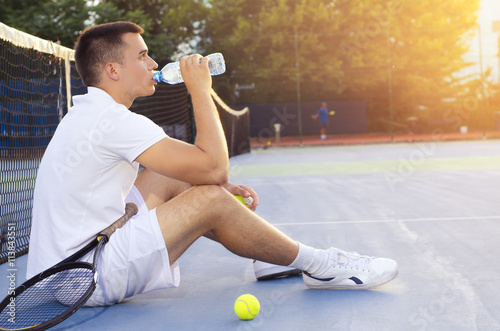 Photography Young tennis player drinking water after playing, sitting on court and looking a