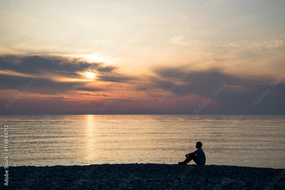 Young man sitting on the stone beach at sea sunset background