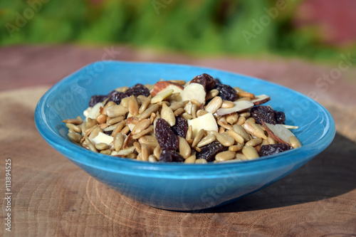 Blue glass bowl filled with trail mix with sliced almonds, salted sunflower kernels, and sugared raisins on a slice of oak wood