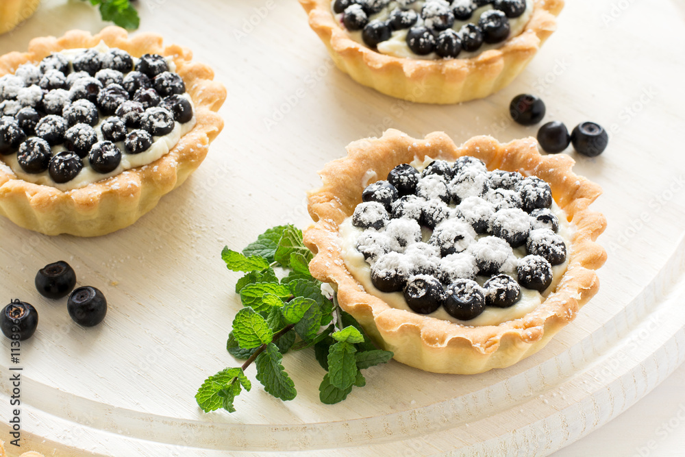 Close up of tartlets with fresh blueberries, decorated mint leaves on white wooden background. Selective focus Close up of tartlets with fresh blueberries, decorated mint leaves on white wooden background. Selective focus