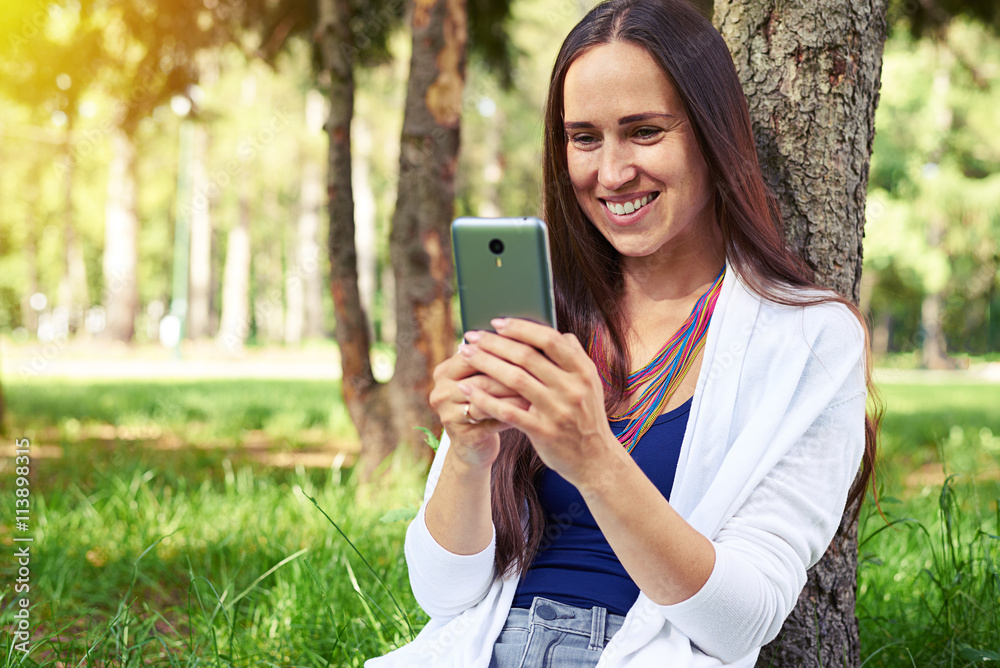Smiling woman sitting under tree and using smart phone Smiling woman sitting under tree and using smart phone