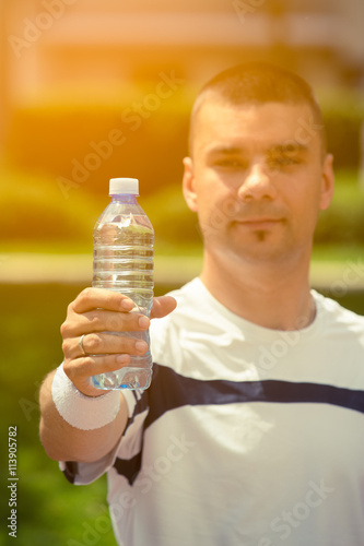Fitness man showing bottle of water in the park. Sunlight and greenery in the background. Ideal for bottle pack shoot adding. Sport, fitness, healthy lifestyle concepts.