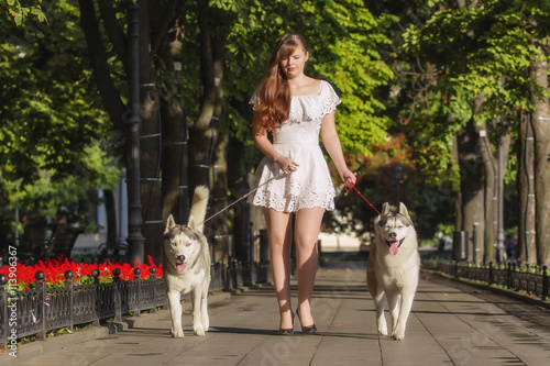 oung girl walking down the street with two dogs. A girl in a white dress. Siberian Huskies.