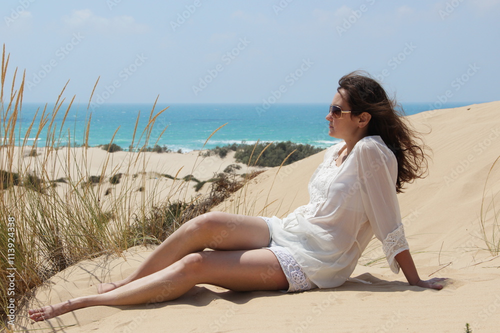 Smiling brunette girl in sunglasses, a white blouse and jeans shorts is sitting on the sand on the background of the desert dunes and the sea Smiling brunette girl in sunglasses, a white blouse and jeans shorts is sitting on the sand on the background of the desert dunes and the sea